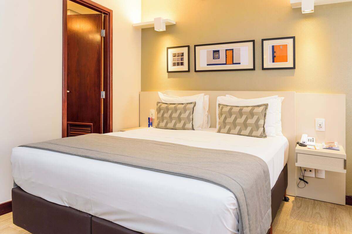 Een hotelkamer met een groot bed, een nachtkastje met een telefoon en een lamp, en lijstjes aan de muur.