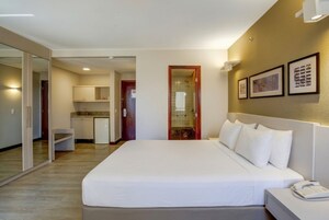 Una habitación de hotel con una cama, una mesita de noche con un teléfono, un espejo, una puerta que da al baño y una kitchenette al fondo.