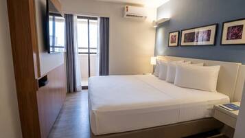 A hotel room with a bed, a desk, a chair, a wardrobe, a window with curtains, and a wall-mounted air conditioner.