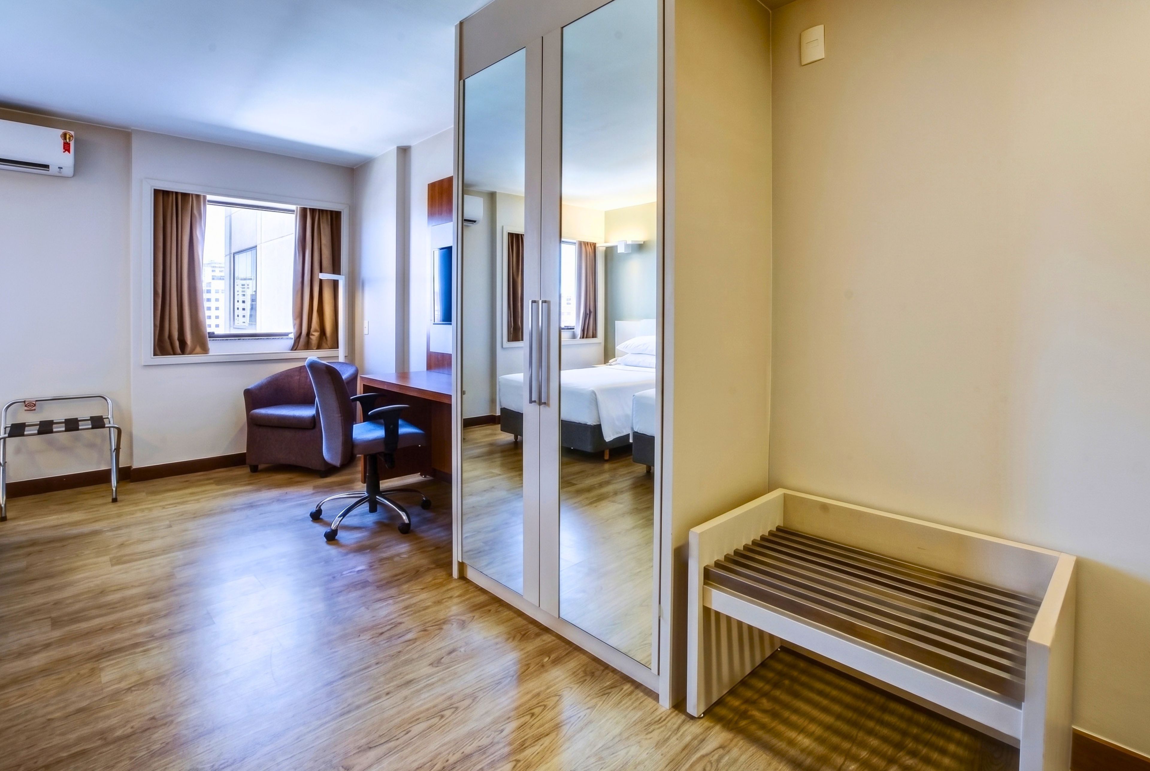 Quarto de hotel com piso de madeira, uma escrivaninha com cadeira, uma cama, uma janela com cortinas e um radiador.