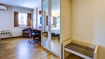Quarto de hotel com piso de madeira, uma escrivaninha com cadeira, uma cama, uma janela com cortinas e um radiador.