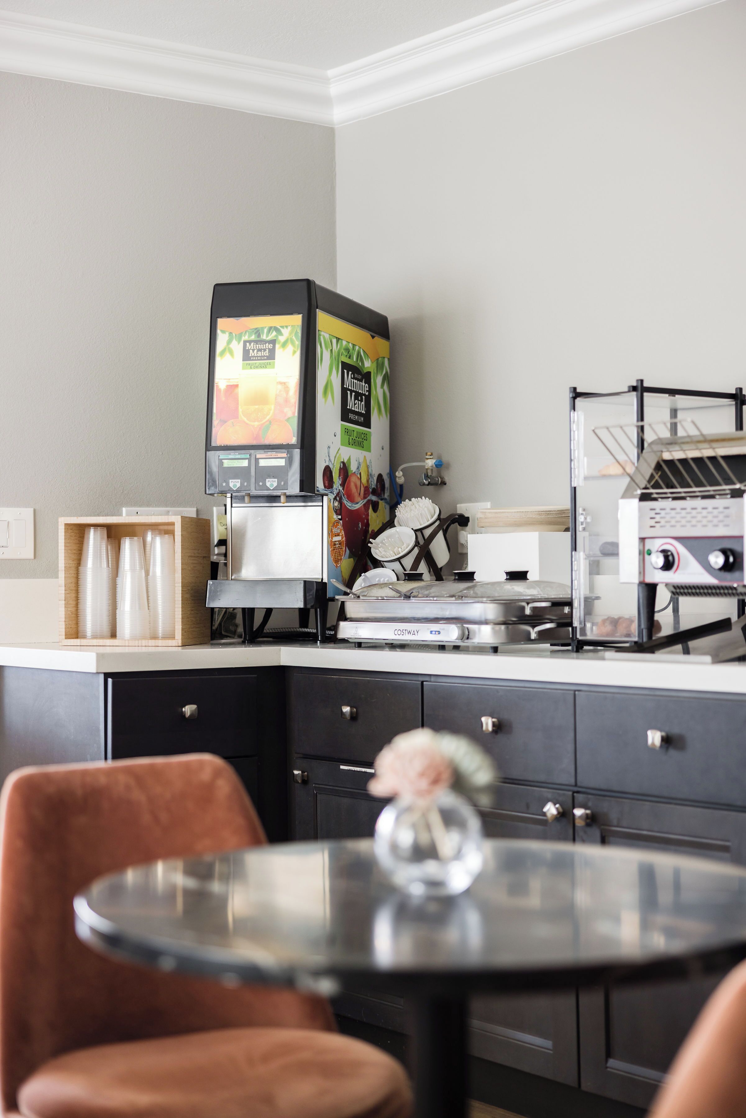 Una cocina moderna con cafetera, microondas y tostadora. Se observan armarios de madera y una pequeña mesa con un jarrón.