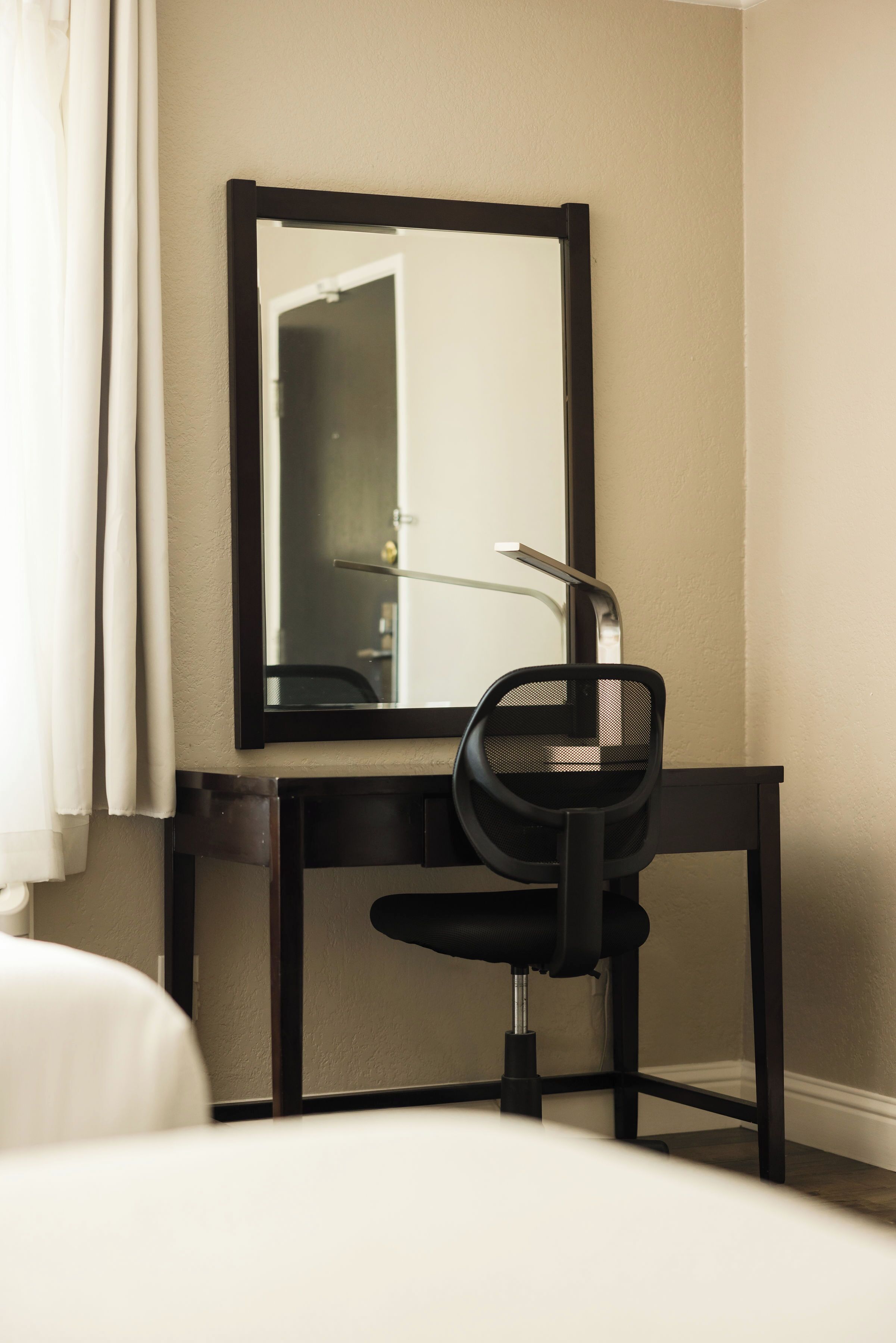 A hotel room with a bed, a desk with a chair, a large mirror, and a window with curtains.