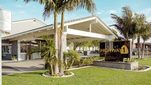 Entrada de hotel con área de estacionamiento techada, palmeras y un letrero que indica 'Highway 1'.