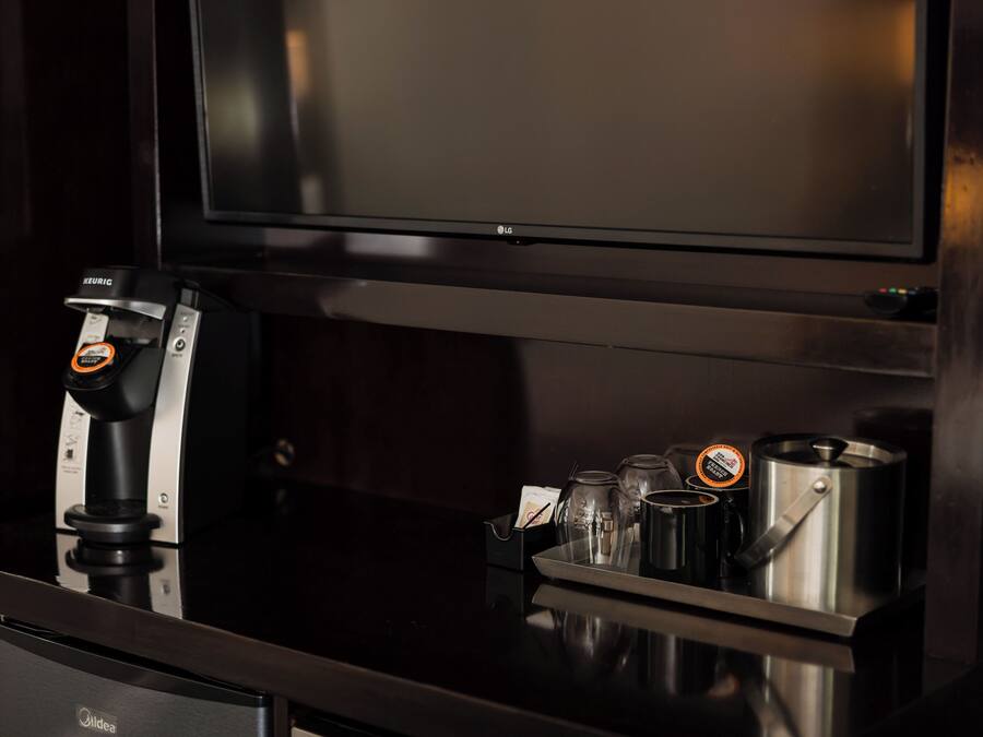 Una cafetera con un cápsula de café Keurig, una jarra de vidrio, una taza y una olla de acero inoxidable sobre una encimera negra reflectante.