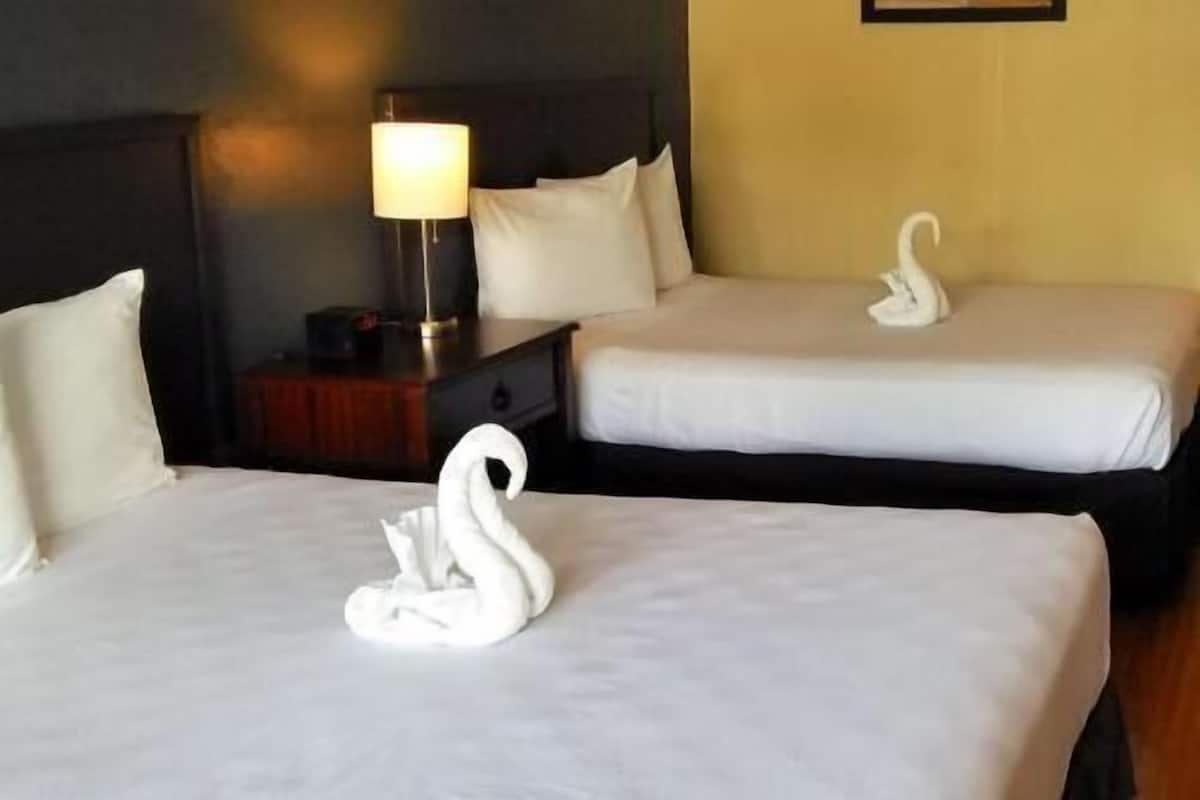 Two beds with white linens, a bedside table with a lamp, and a wall-mounted picture frame.