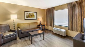A hotel room with a sofa, armchair, coffee table, two lamps, a framed picture, a large window with curtains, and an air conditioning unit.
