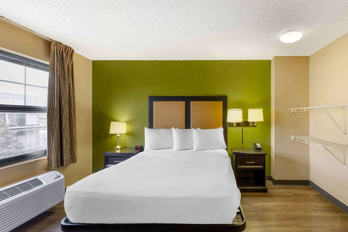 A hotel room with a large bed, two bedside tables with lamps, a window with curtains, a wall-mounted heater, and a wall-mounted drying rack.