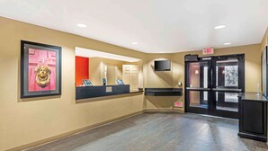 A hotel reception area with a reception desk, a digital check-in kiosk, a television mounted on the wall, and an exit sign.