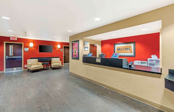 Zona de recepción de un hotel con pared roja, un cartel que dice 'WELCOME', mostrador de recepción y dos sillones.