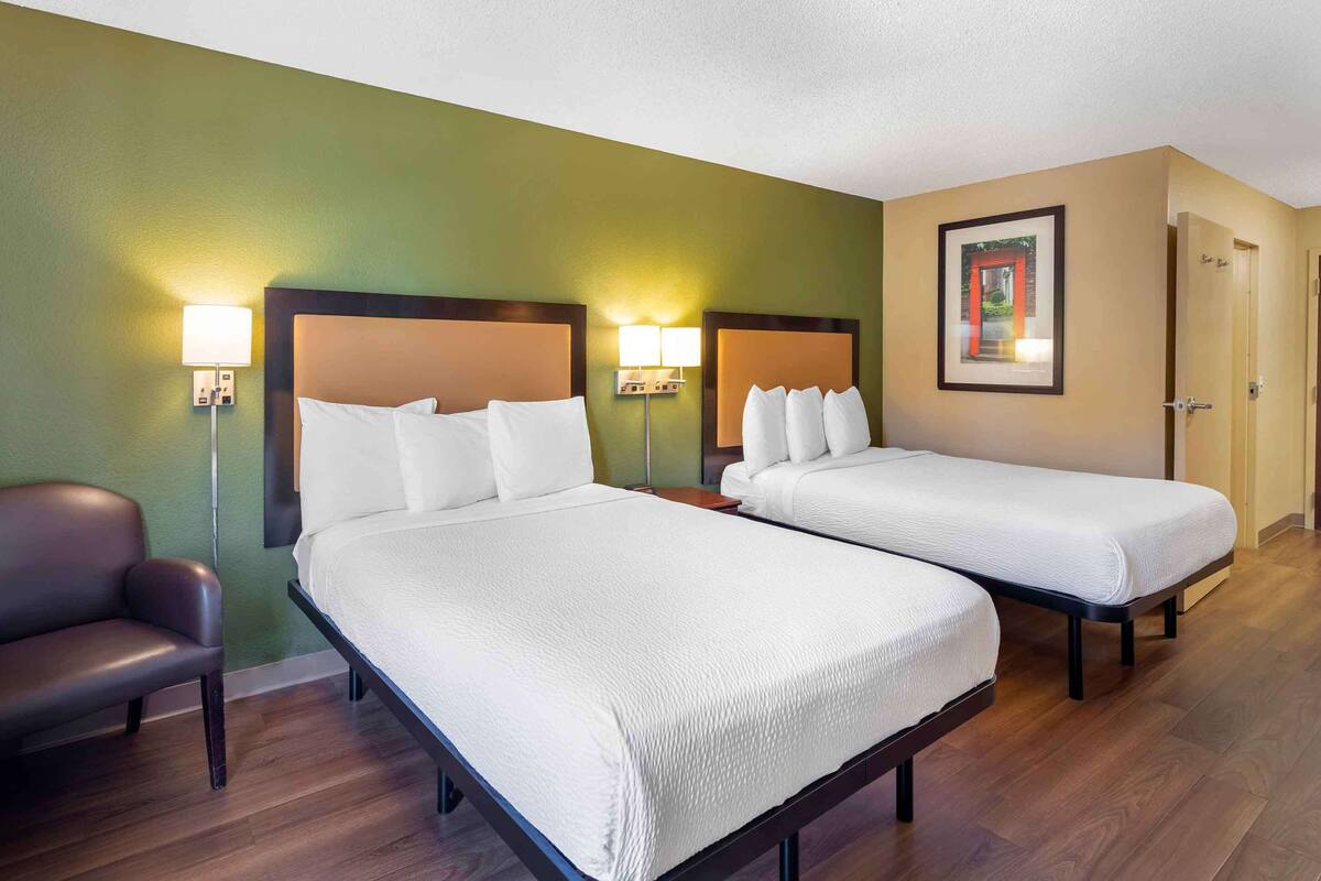 A hotel room with two beds, a chair, and wall-mounted lamps.