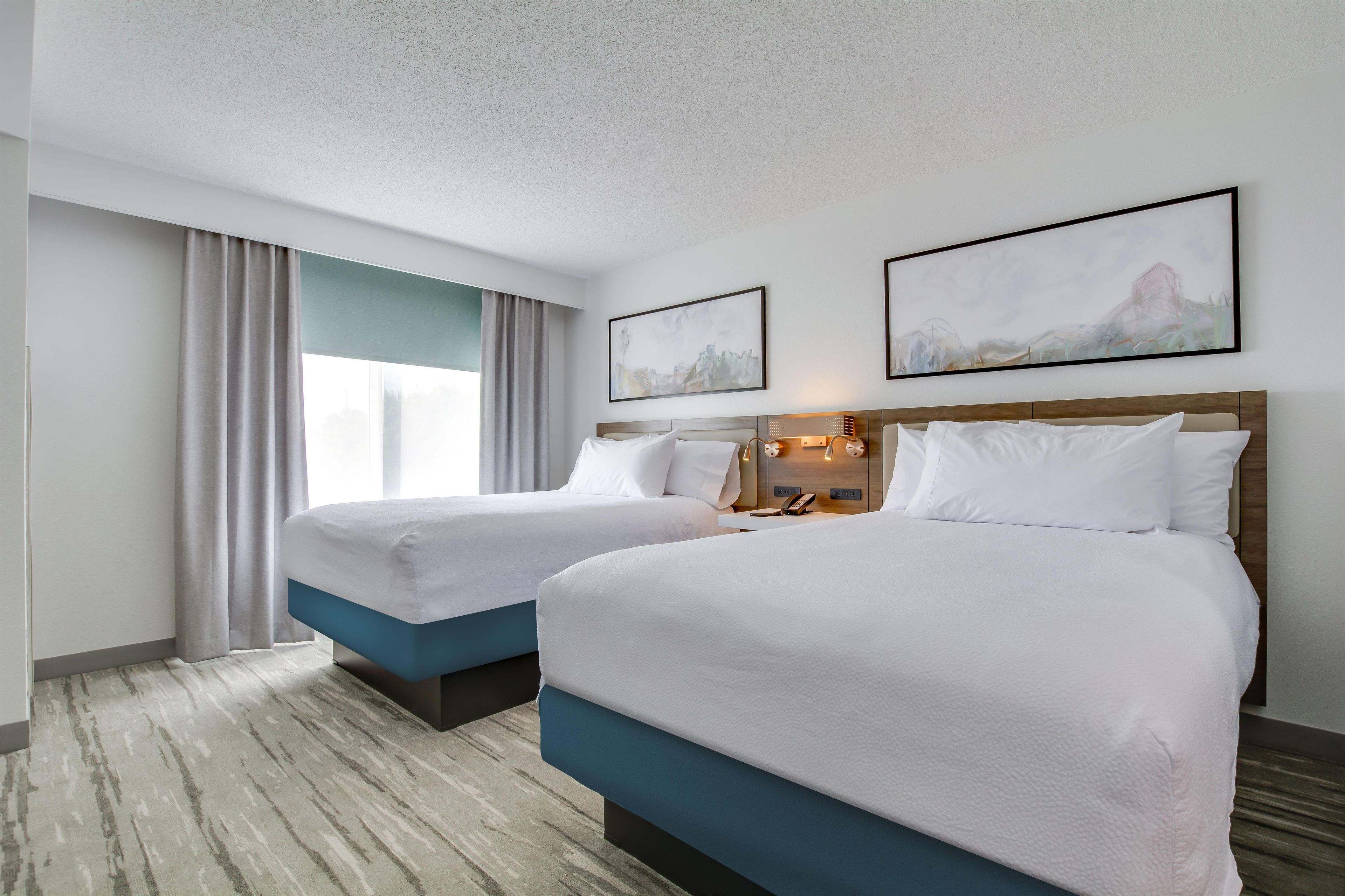 A hotel room with two beds, a large window with curtains, a wall-mounted phone, and abstract art.
