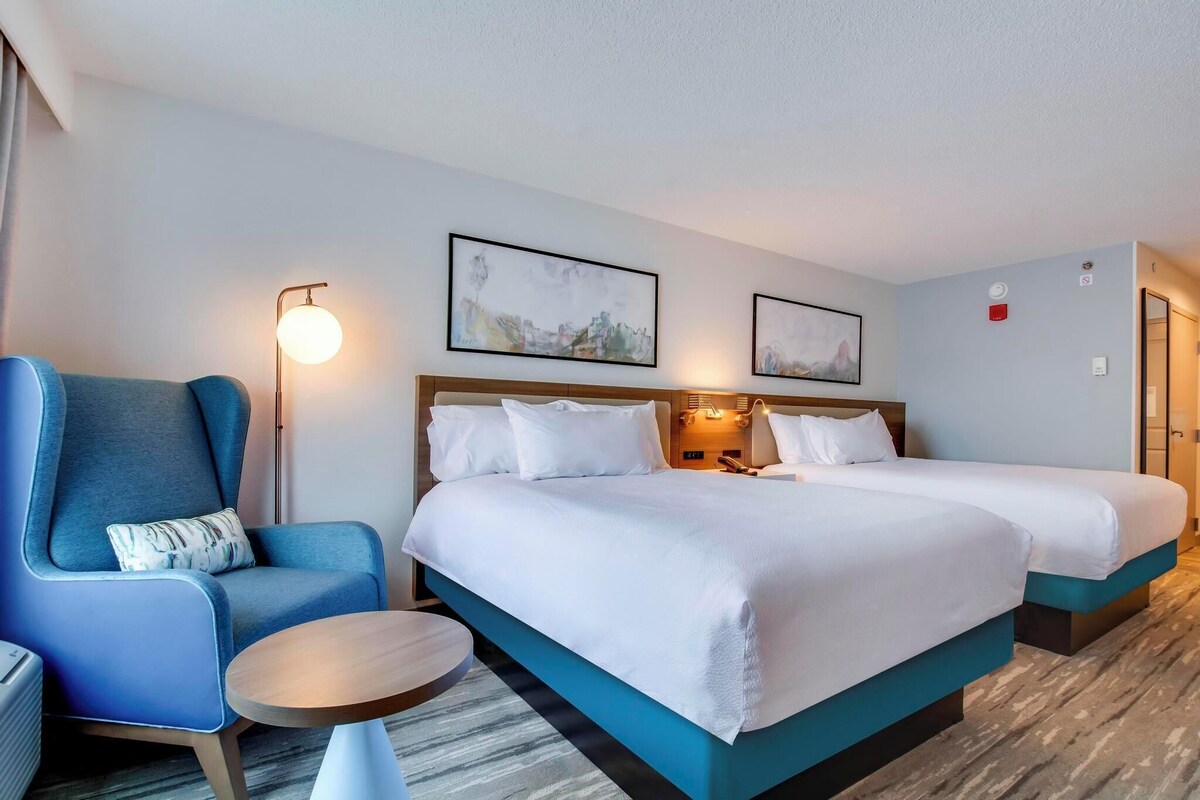 A hotel room with two beds, a blue armchair, a round table, and artwork on the walls.