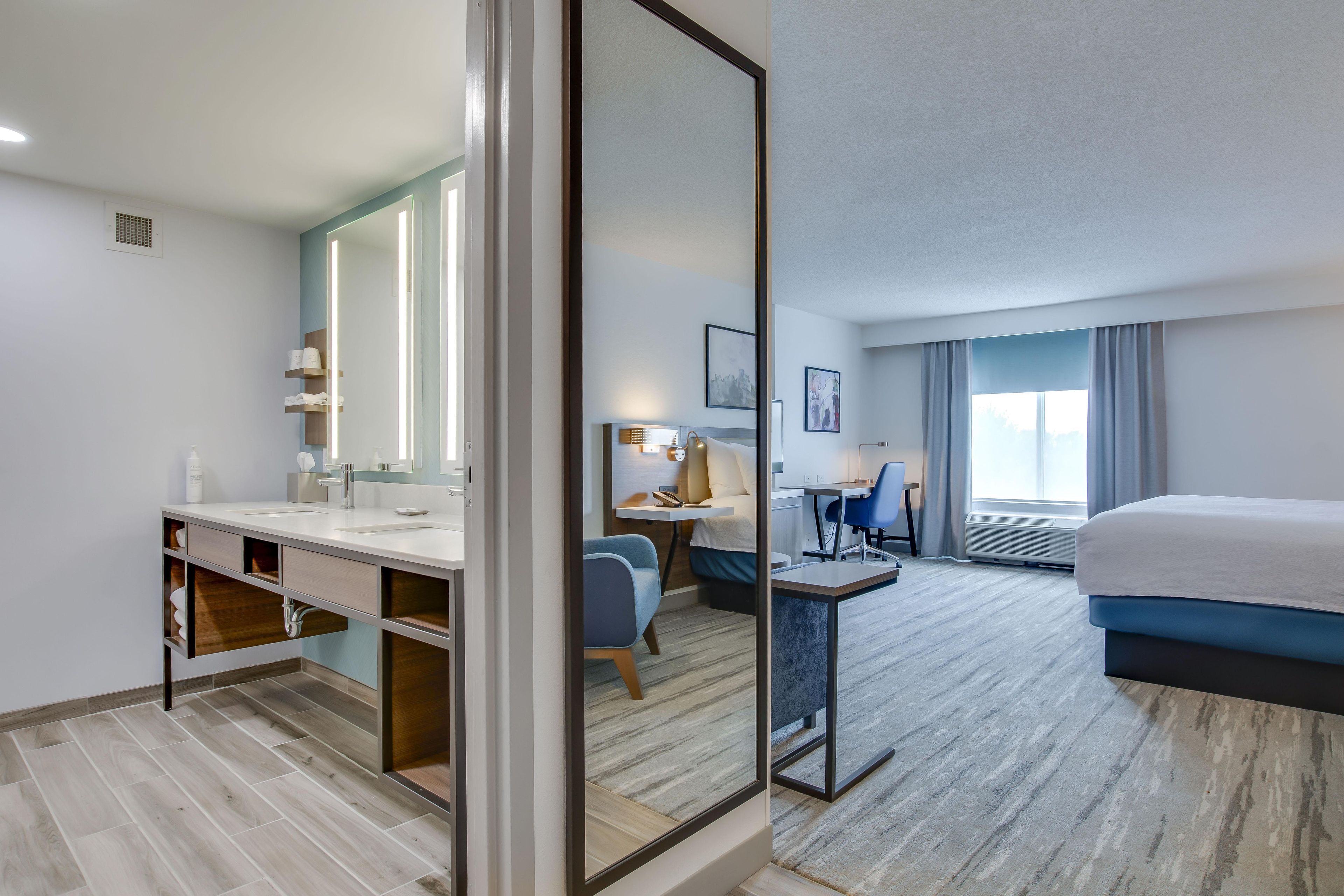 Habitación de hotel con cama, escritorio, silla, espejo y baño con lavabo y toallero.