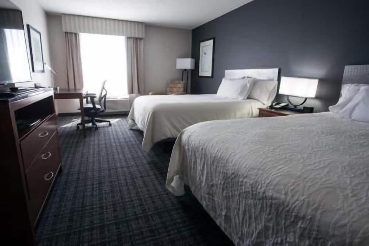 A hotel room with two beds, a desk, a chair, a lamp, and a window with curtains.