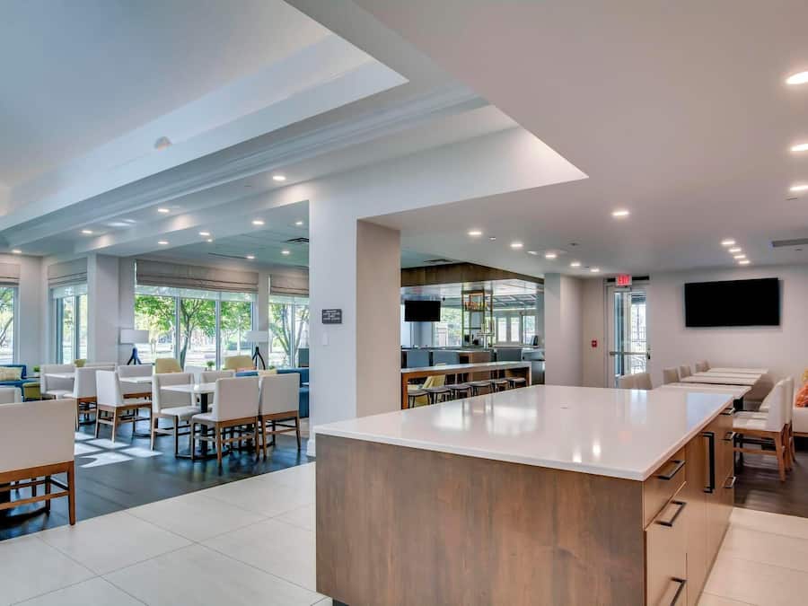A modern dining area with a long counter and multiple tables, large windows, and a flat-screen TV mounted on the wall.