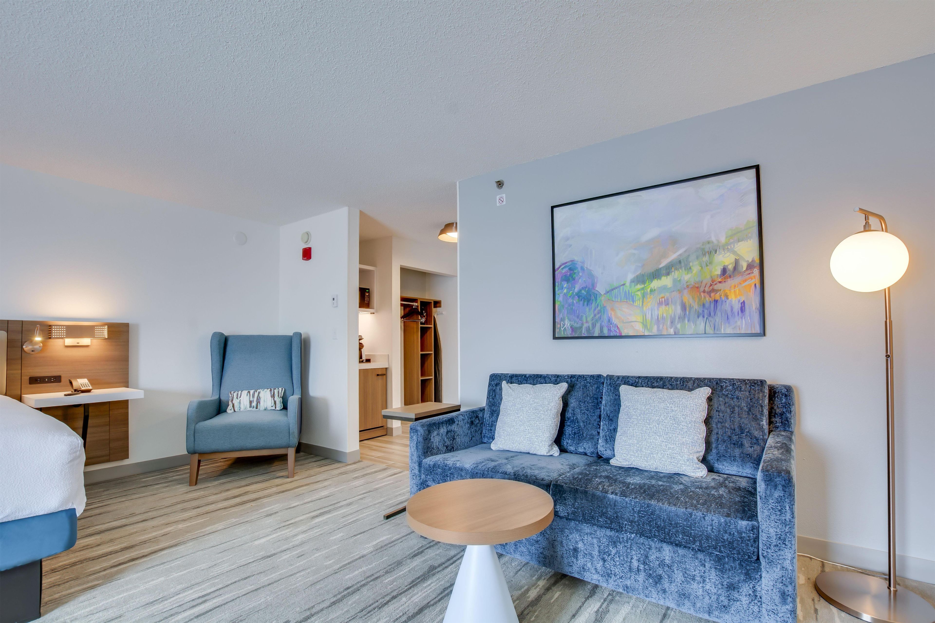 A modern hotel room with a sofa, a small table, a chair, a bed, a desk, and a painting on the wall.