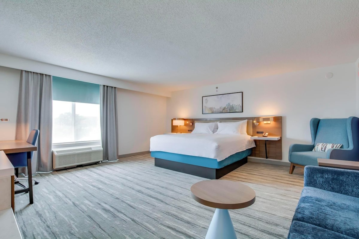 A hotel room with a bed, a desk, a chair, a small table, and a window with curtains.