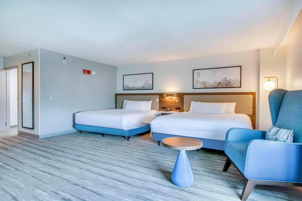 A hotel room with two beds, a small round table, and a blue armchair.