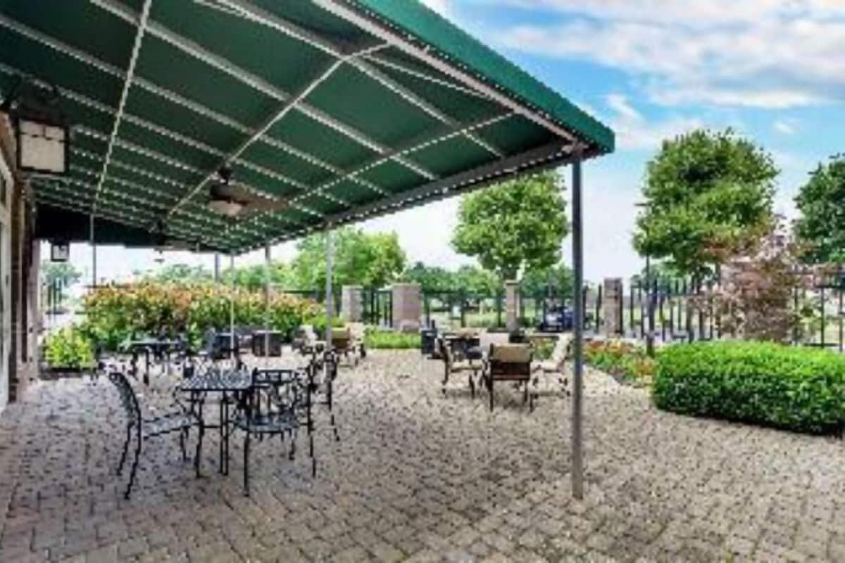 A covered outdoor seating area with tables and chairs, surrounded by greenery and trees.