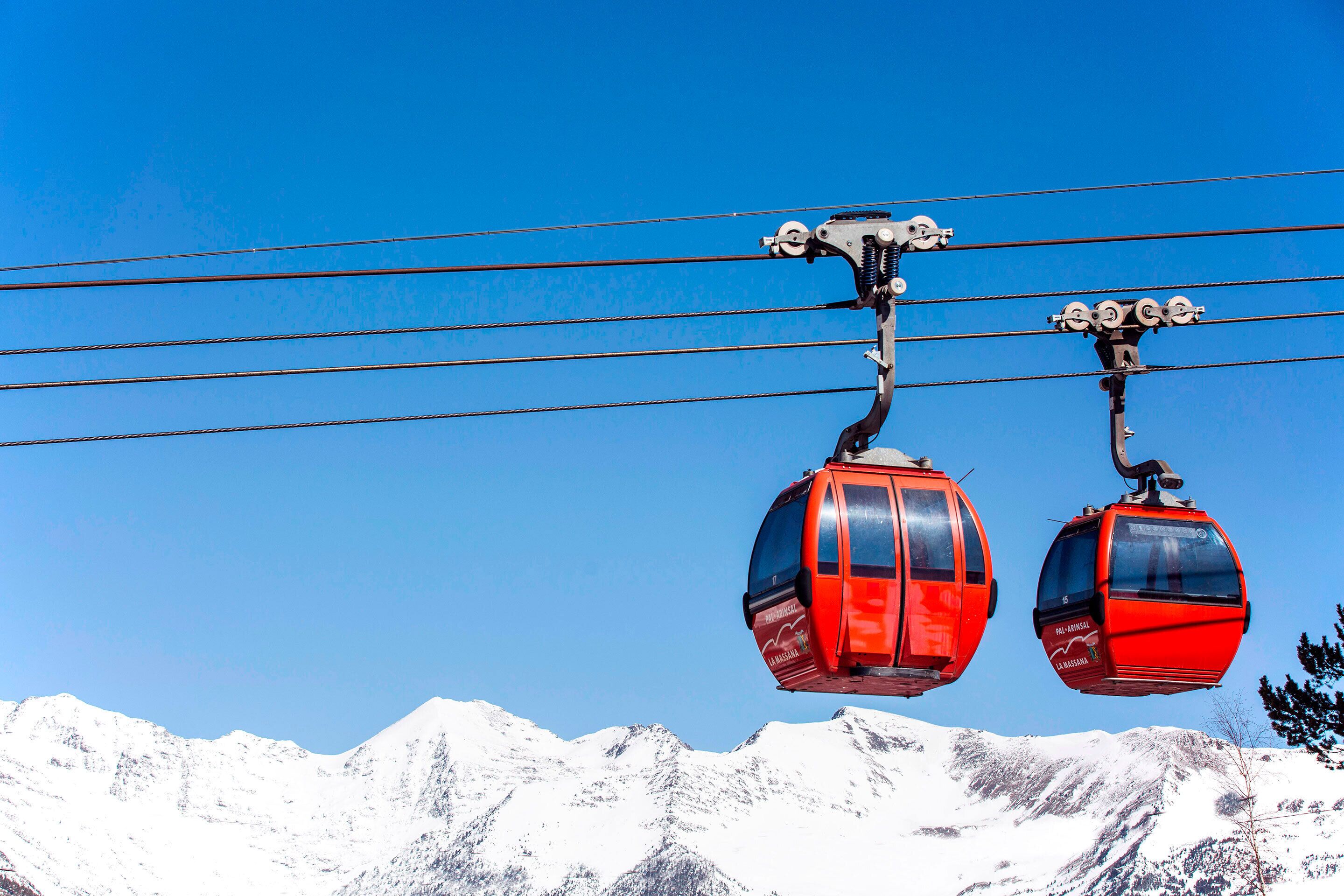 Two red cable cars suspended by a cable system against a clear blue sky, with snow-covered mountains in the background.