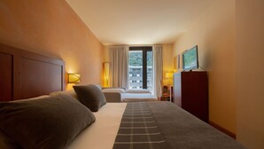 A hotel room with a large bed, two bedside tables with lamps, a TV on a stand, and a view of a building through the window.
