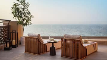 Un balcón con mobiliario de mimbre, una pequeña mesa con un florero y fruta, y vista al mar.