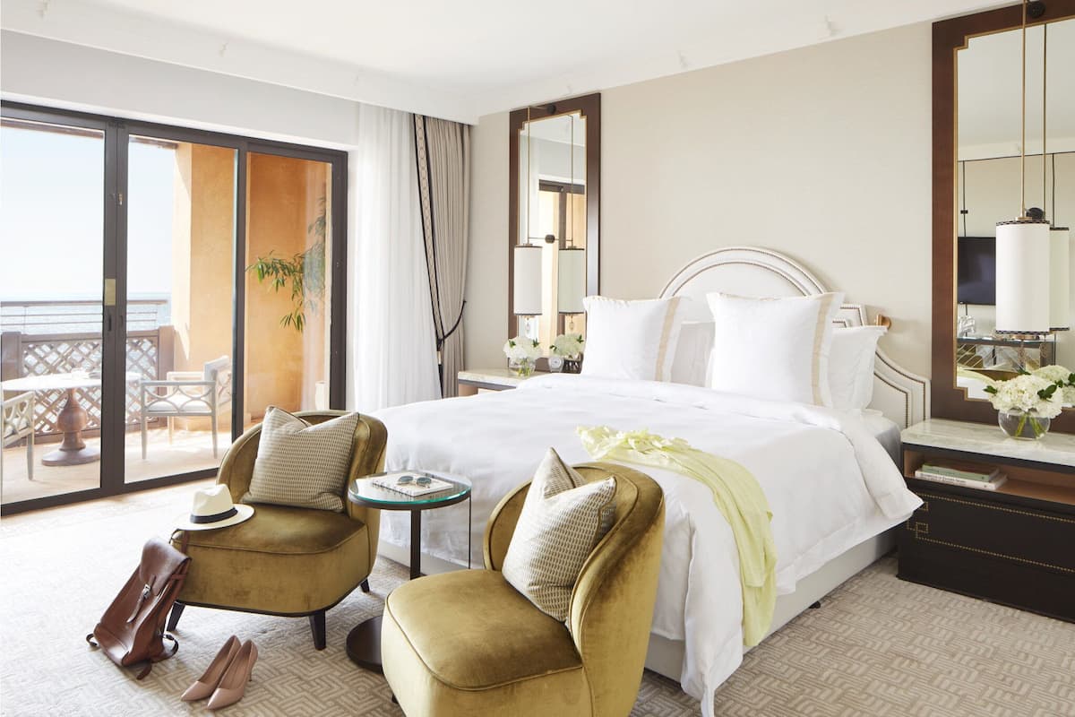 A hotel room with a large bed, two armchairs, a small table, and a view of the outdoors.