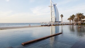 A luxurious infinity pool with a view of the sea and a distinctive sail-shaped building in the background.
