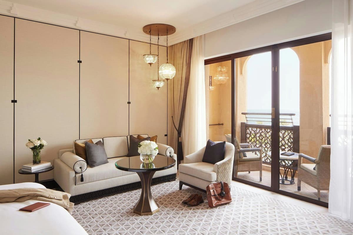 A hotel room with a sofa, armchair, and a table with a vase of flowers. There is a balcony with outdoor furniture.