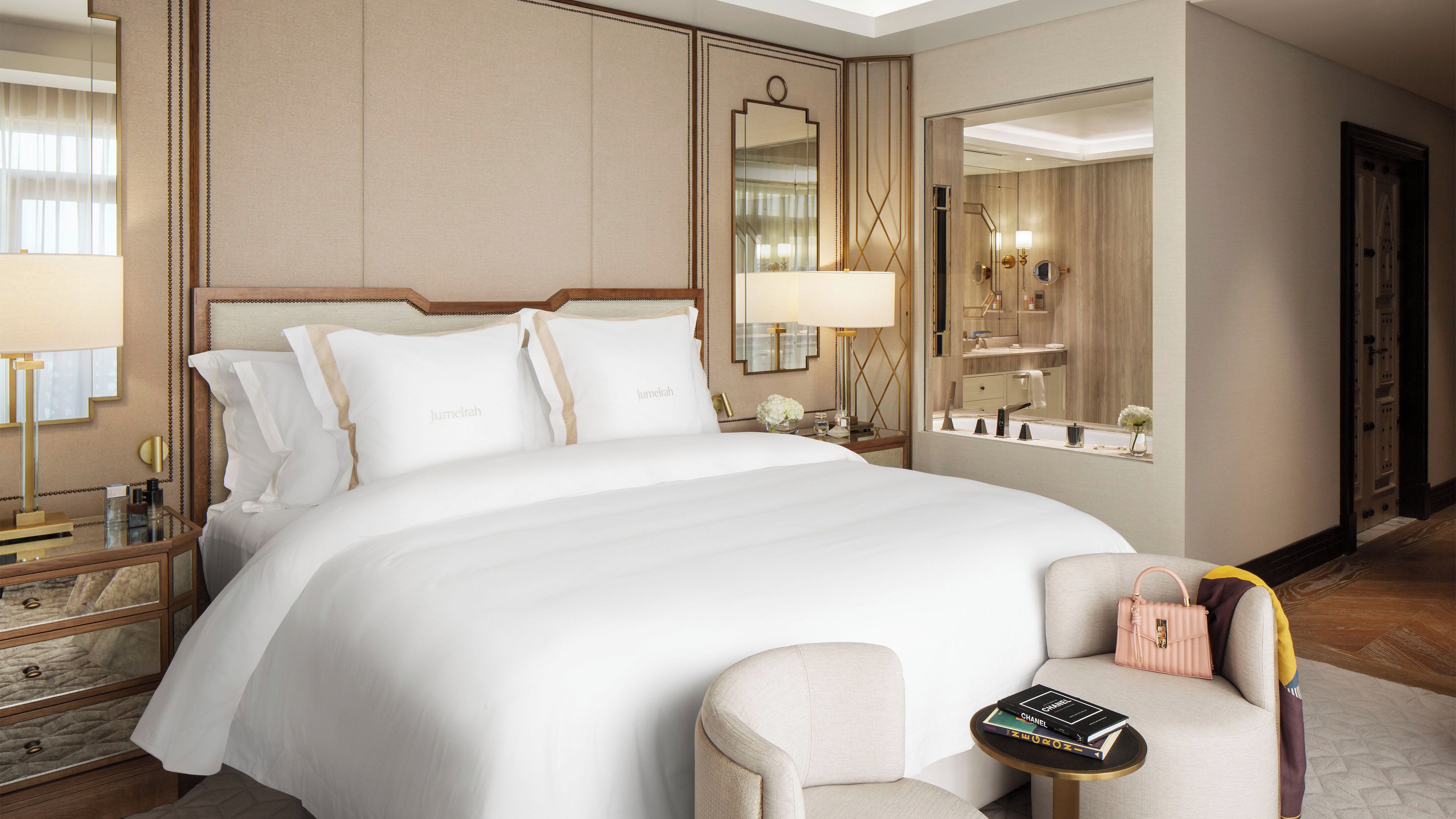 A hotel room with a large bed, bedside tables, a mirror, a chair, and a small table with books.
