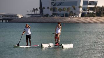 Two people on paddleboards in a calm body of water with a modern building and palm trees in the background.