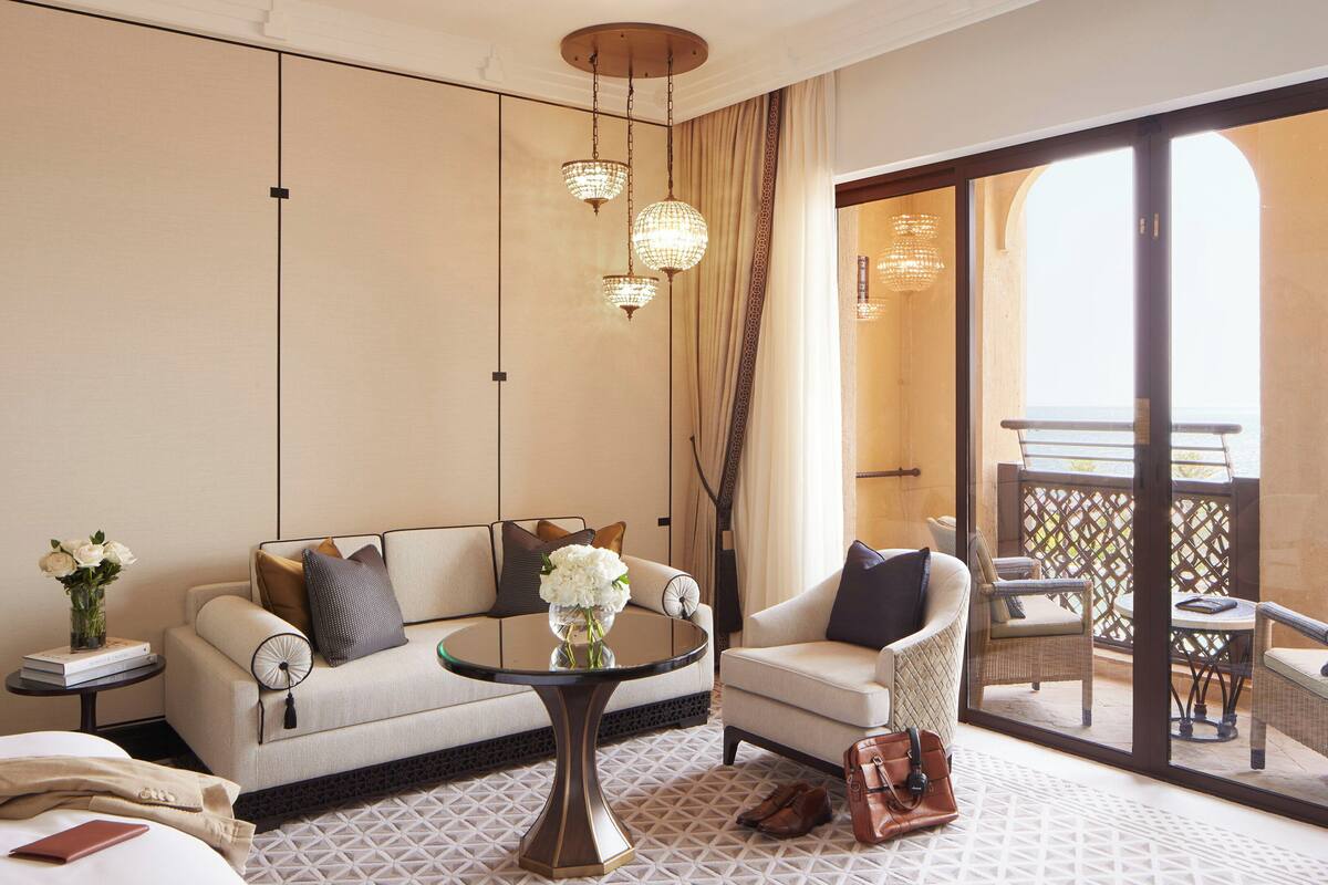 A hotel room with a sofa, armchair, round table, and a view of the balcony with outdoor furniture.