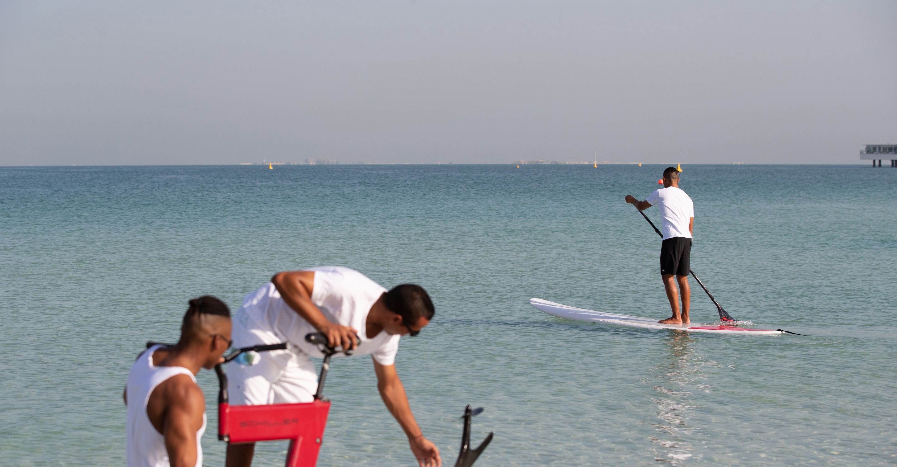 Una persona haciendo paddleboard en un mar tranquilo, con una costa lejana y un muelle.