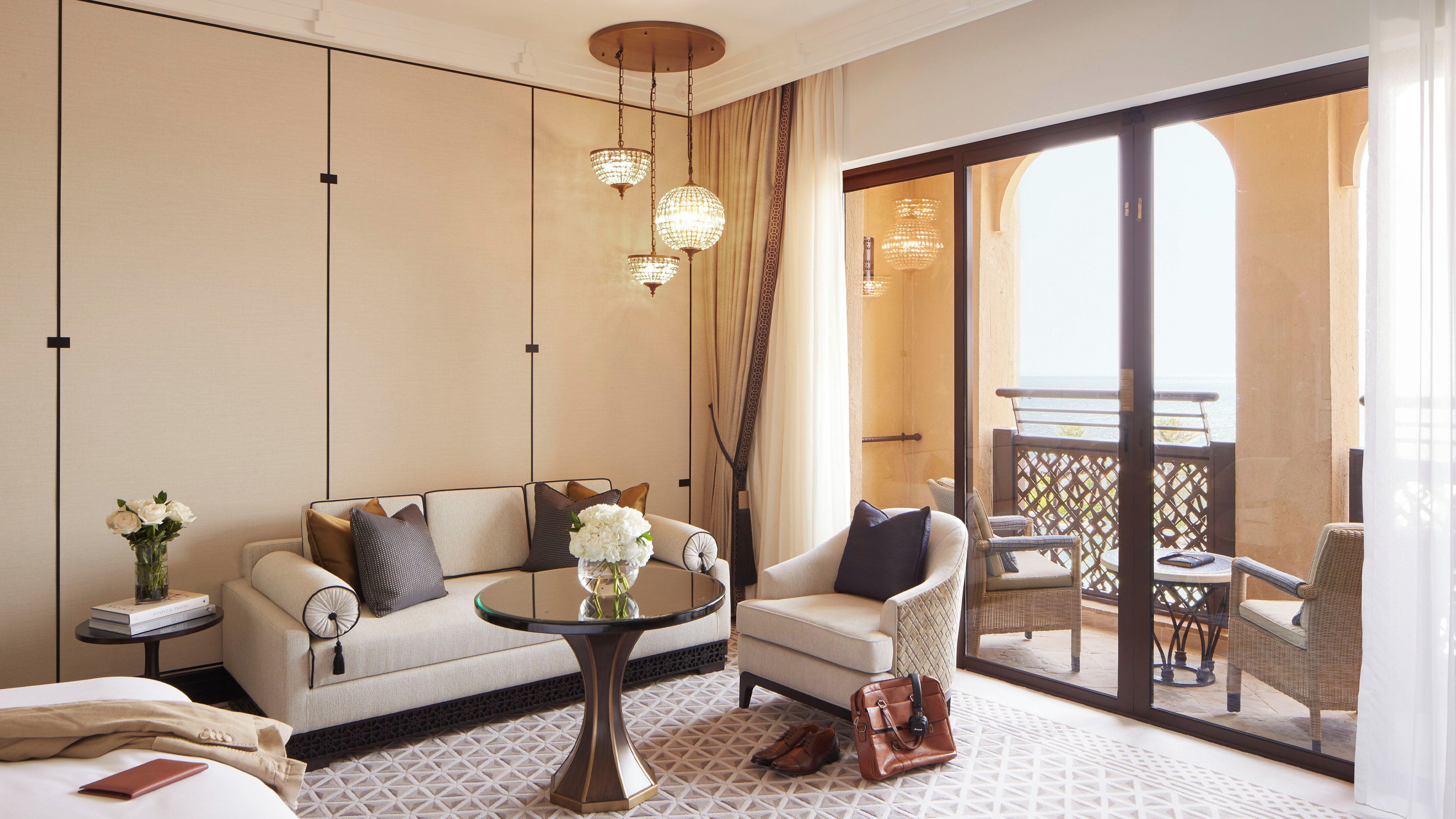 A hotel room with a sofa, armchair, round table, and a balcony with wicker furniture.