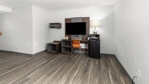 A hotel room with a flat-screen TV, a desk with a lamp, a small refrigerator, and a chair.