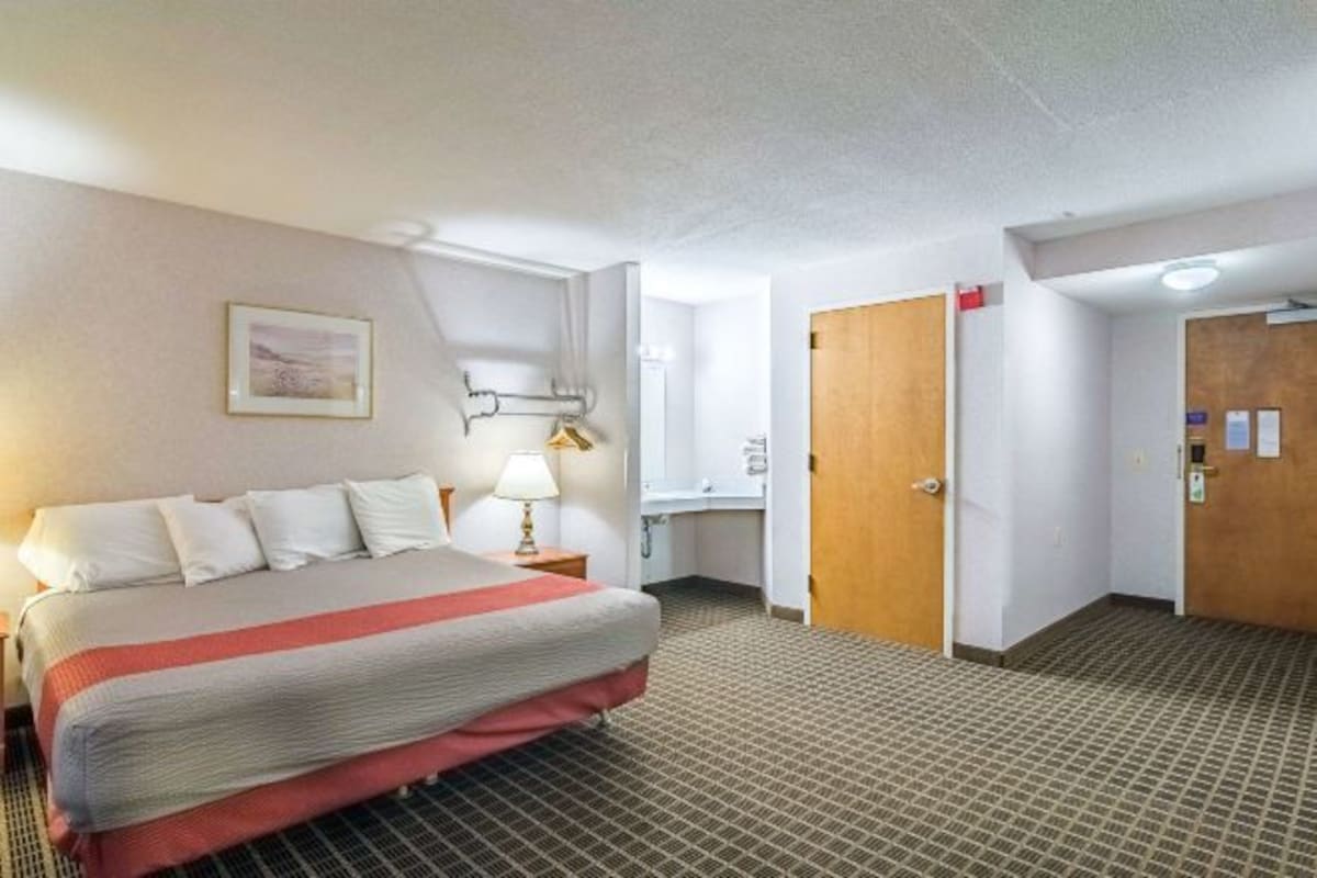 A hotel room with a bed, a desk, a chair, a lamp, a door, and a wall-mounted light fixture.