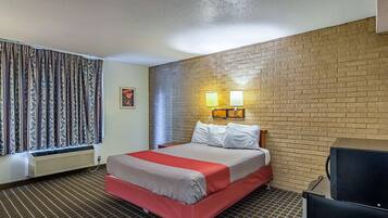 Una habitación de hotel con cama, un calentador, una ventana con cortinas, una pared de ladrillo y una lámpara fijada a la pared.