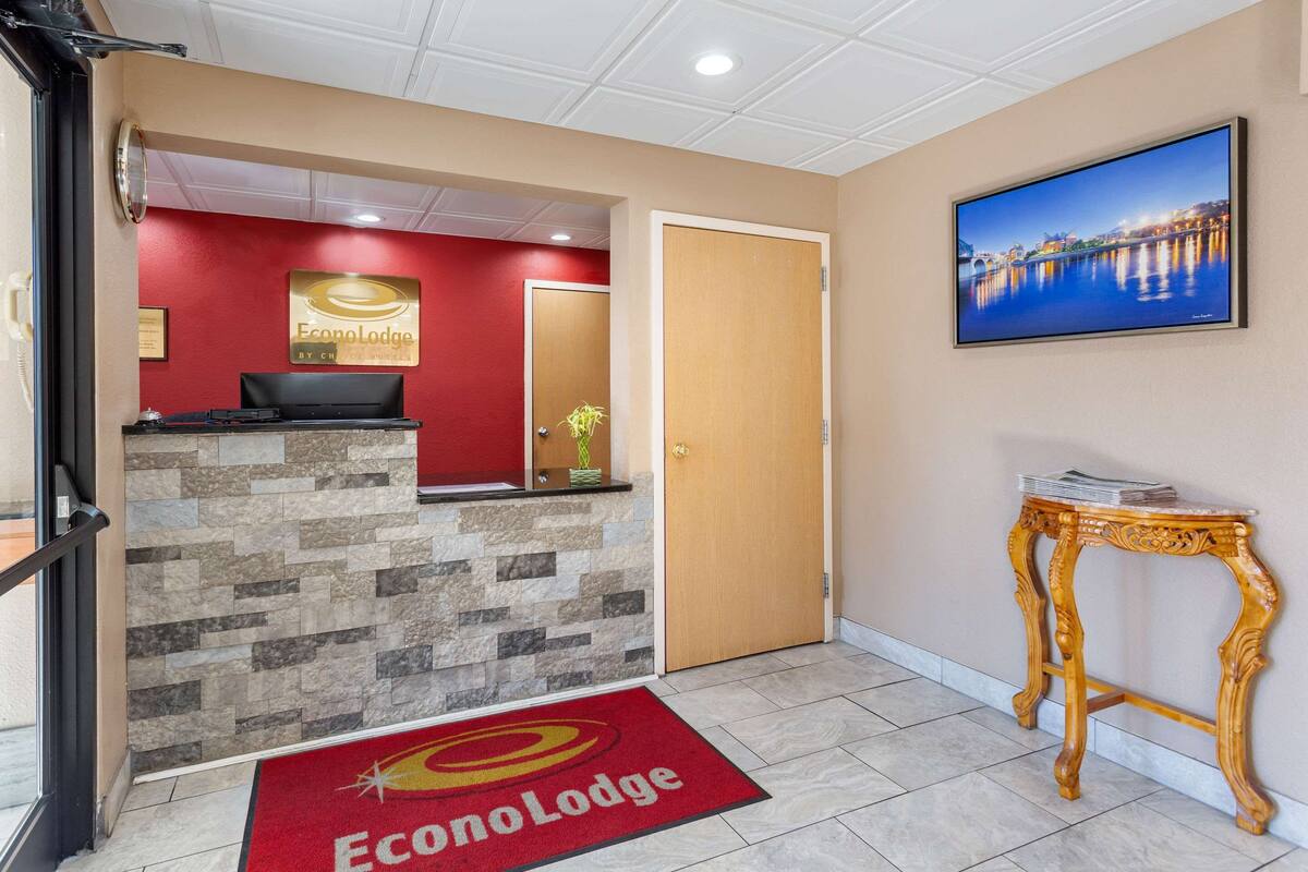 A hotel reception area with a stone counter, a red carpet with the hotel logo, a wooden door, and a wall-mounted TV displaying a scenic view.