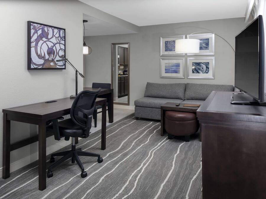 Habitación de hotel moderna con escritorio, silla de oficina, sofá y un televisor de pantalla plana grande.