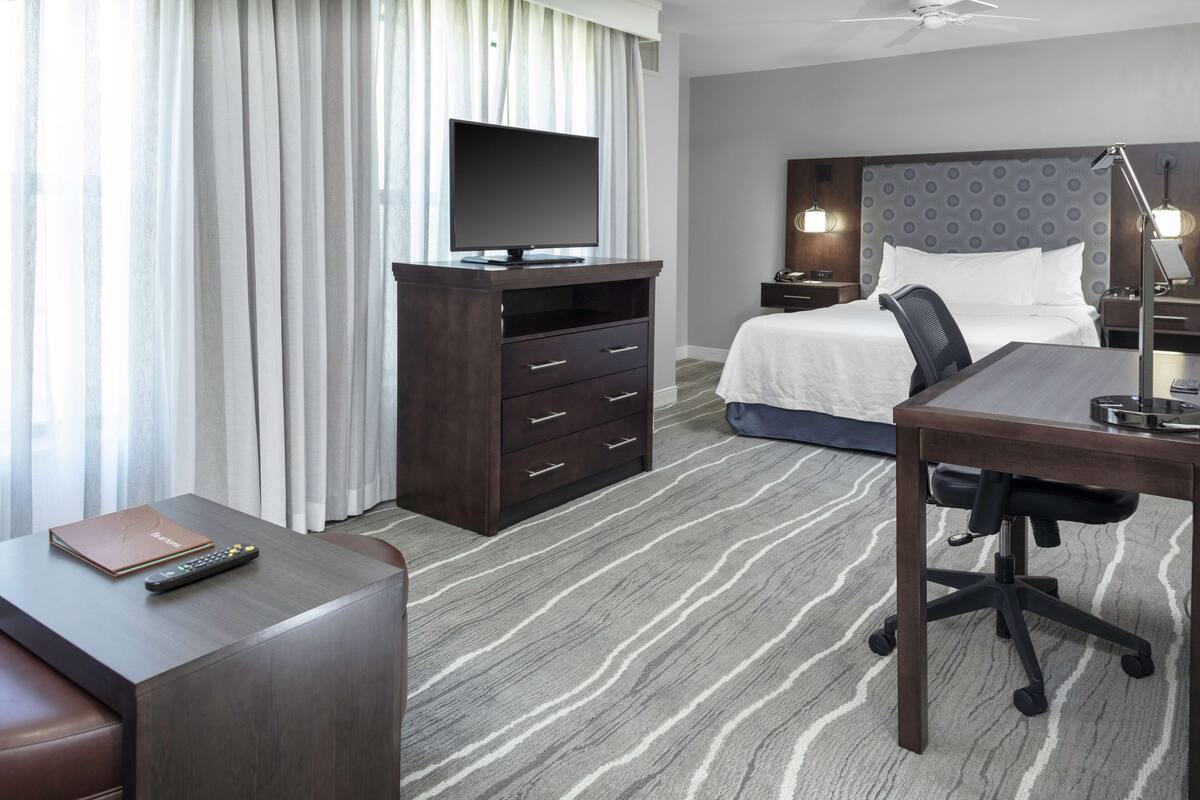 A hotel room with a bed, desk, chair, TV, dresser, and curtains.