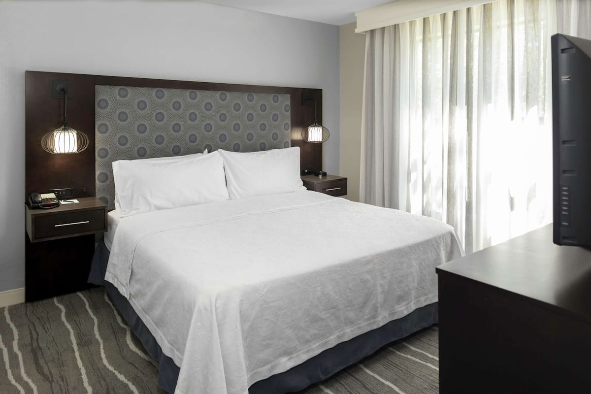 A hotel room with a large bed, two bedside tables, a TV, and a window with curtains.