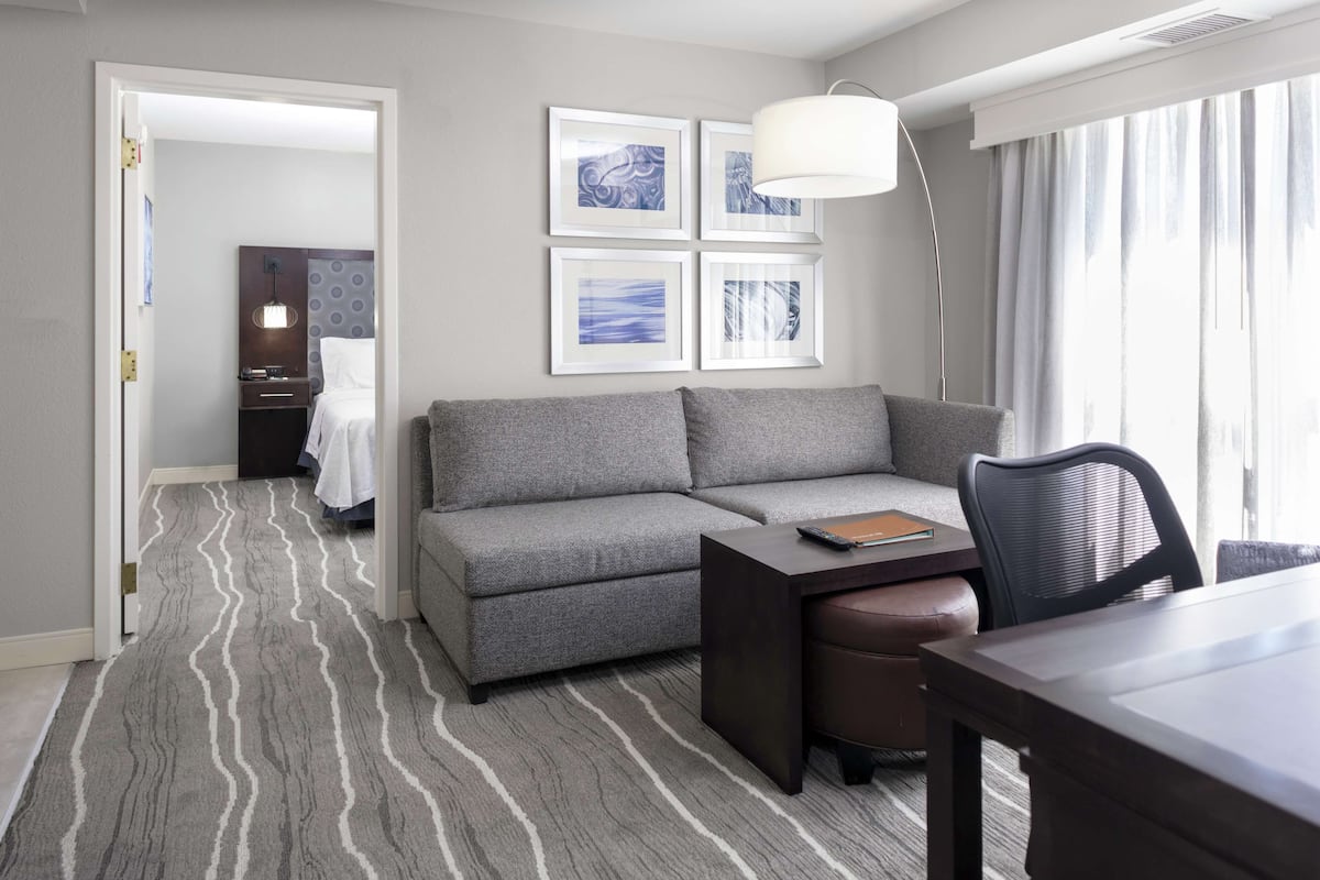 A modern hotel room with a gray sofa, a small table with a remote control, a desk, and a chair. There are framed pictures on the wall and a large window with curtains.