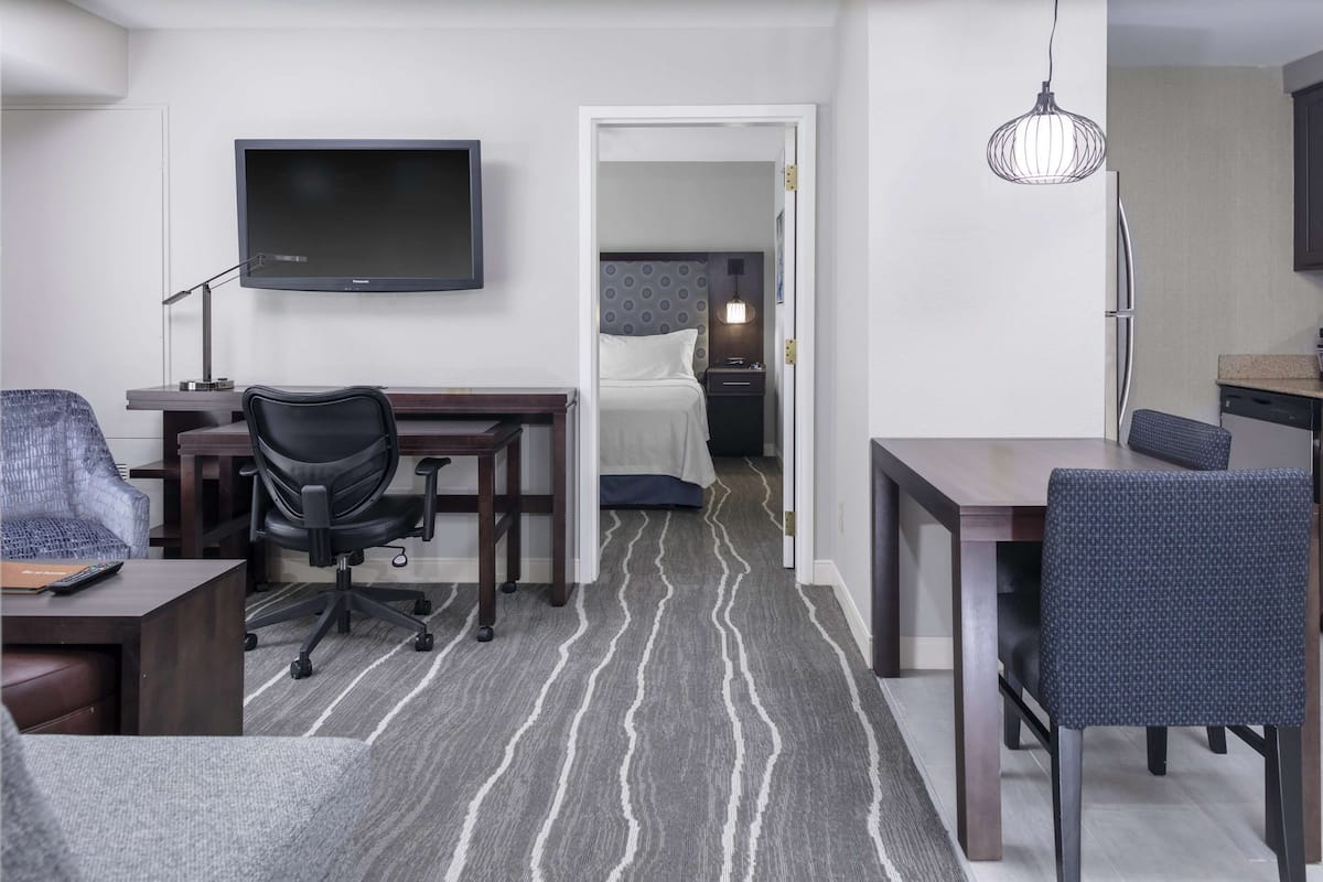 A hotel room with a flat-screen TV, a desk with a chair, a dining table with a chair, a bed with a headboard, and a nightstand with a lamp.