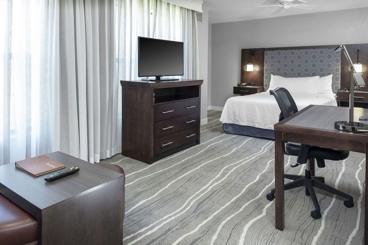A hotel room with a bed, a desk, a chair, a TV on a stand, a dresser, and a window with curtains.
