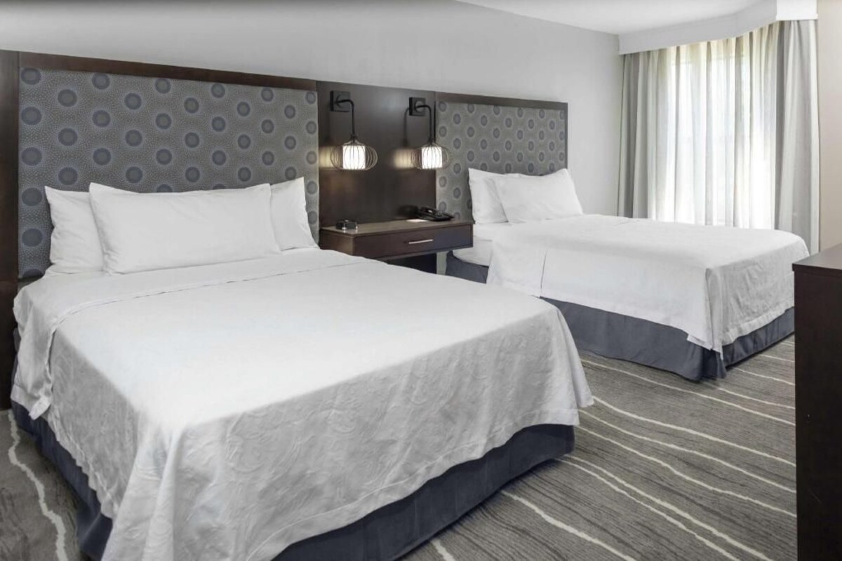 A hotel room with two beds, a headboard with a circular pattern, bedside lamps, a nightstand, and a large window with curtains.