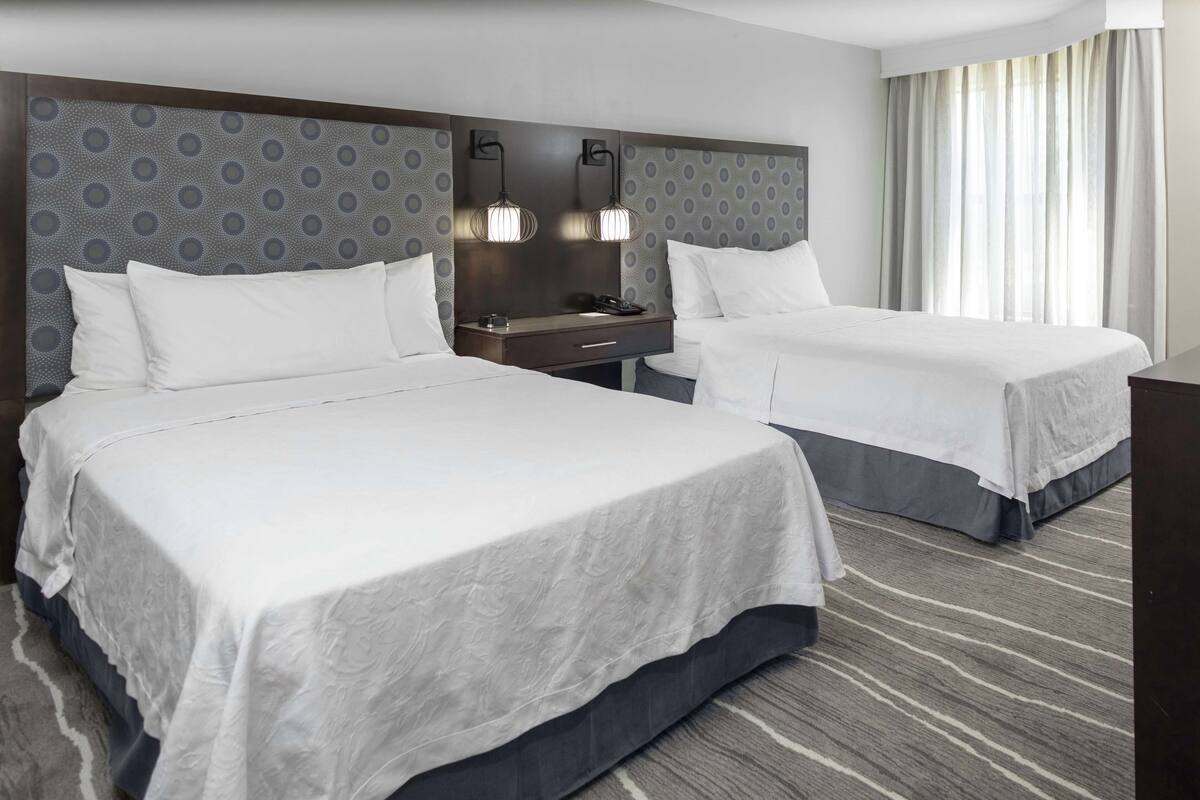A hotel room with two beds, a nightstand, a desk, and a large window with curtains.