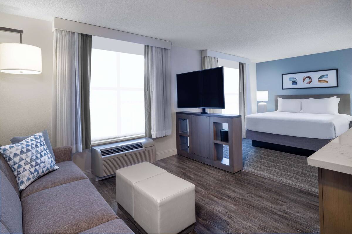 A hotel room with a sofa, ottoman, flat-screen TV, bed, and artwork on the wall.