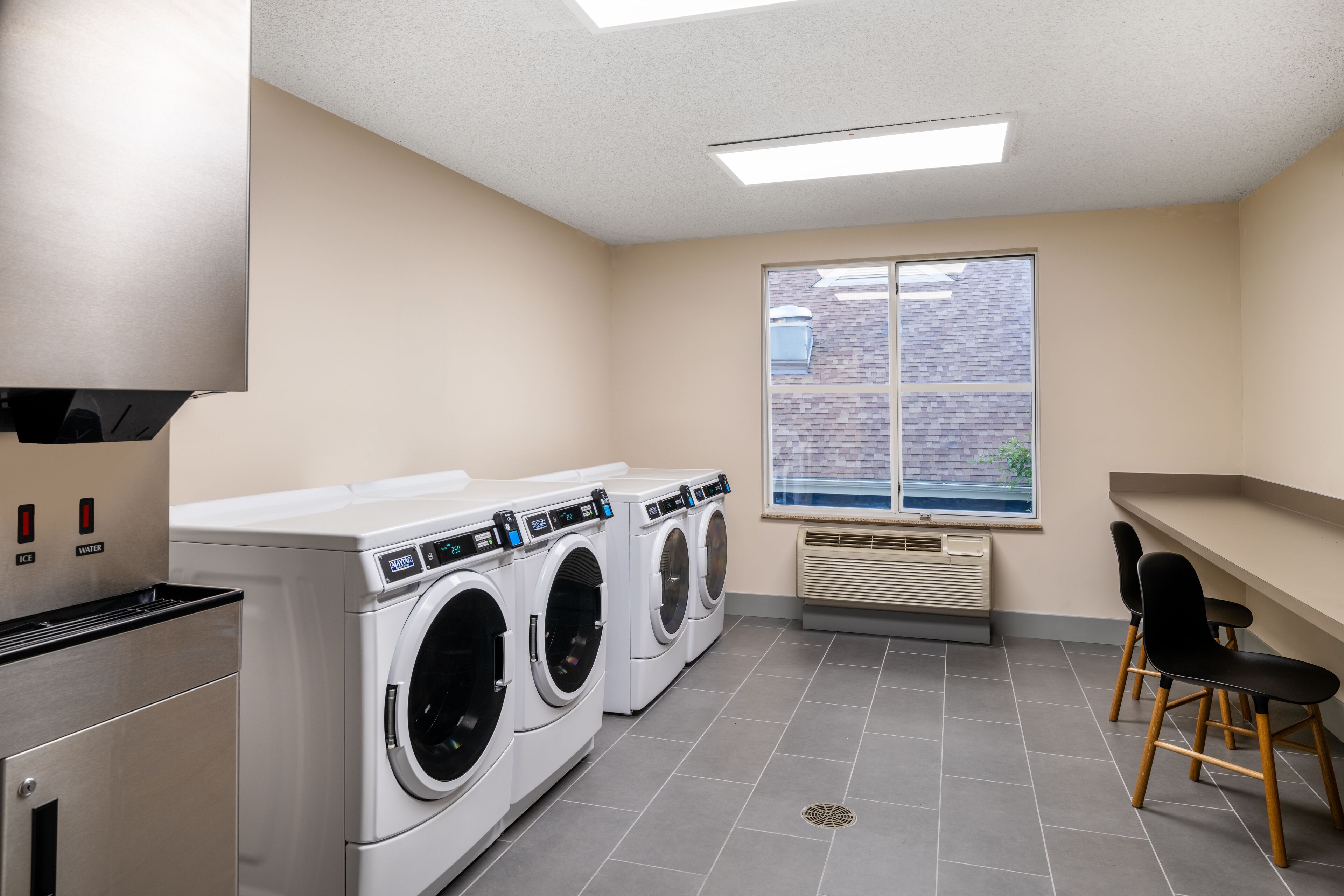 A laundry room with three front-loading washers and dryers, a ventilation unit, and a window with a view of a brick building.