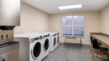 Uma lavanderia com três máquinas de lavar e secar de abertura frontal, uma unidade de ventilação e uma janela com vista para um edifício de tijolos.
